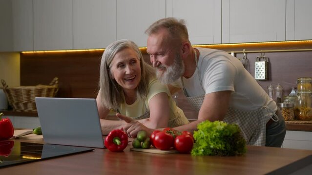 Older Caucasian Couple Middle-aged Spouses Family Using Laptop Device At Home Kitchen Choosing Food Delivery Fresh Vegetables Online Shopping Natural Products Order Retired Woman And Man Laughing Fun