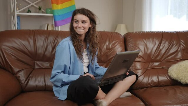 A teenage girl with long hair is taking a video call over the Internet, talking using a laptop, sitting in the lotus position on a leather sofa in a spacious living room with a rainbow flag on the