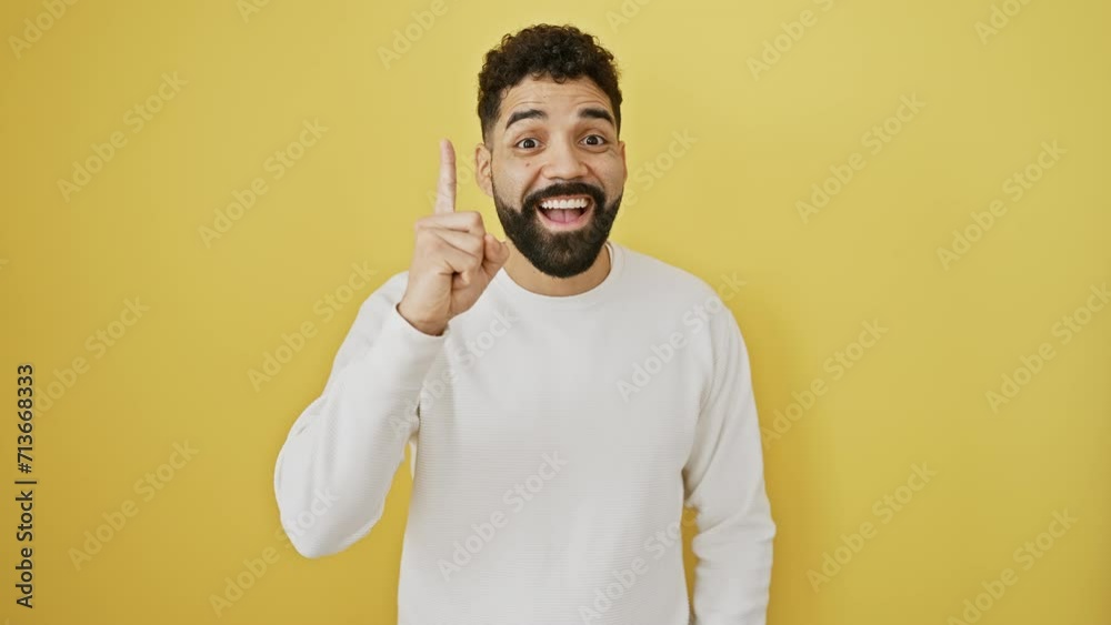 Wall mural Cheerful young hispanic man with a beard, gesturing the number one with his finger, brimming with a clever idea. exuding success and joy against an isolated yellow wall.