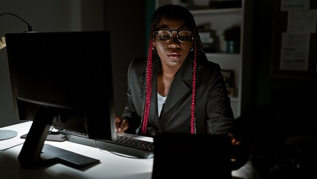 Serious african american woman, a focused business worker, engrossed during her night work at the office, professionally handling her computer and touchpad amidst the room's dim light.