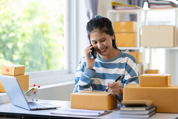 Young Asian woman starting an online selling business at home using mobile phone is checking online messages and calling customers.