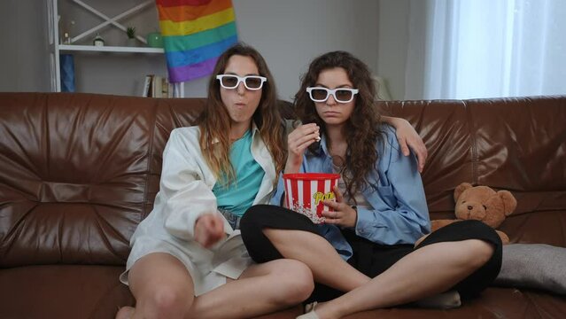 Two girlfriends wearing 3D glasses are watching a horror movie and eating popcorn, while sitting on a leather sofa in the living room. The girl got scared, took off her glasses and complains to her