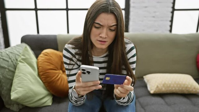 Hispanic teenager holding a credit card and smartphone looks worried while sitting on a sofa indoors, depicting financial concern.