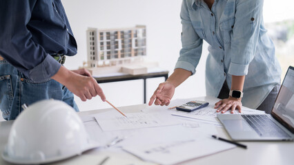 Two engineer architect pointing on blueprint of sketching interior architectural building to discussion technical for construction plan and building model while working together in workplace site
