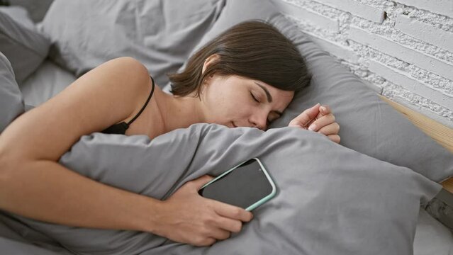 Beautiful young hispanic woman dozing off, lying in bed holding smartphone in cozy bedroom, visibly tired after morning routine.