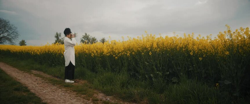 Footage of young lady using a binoculars anf standing on a trail in a rapeseed field on a cloudy day