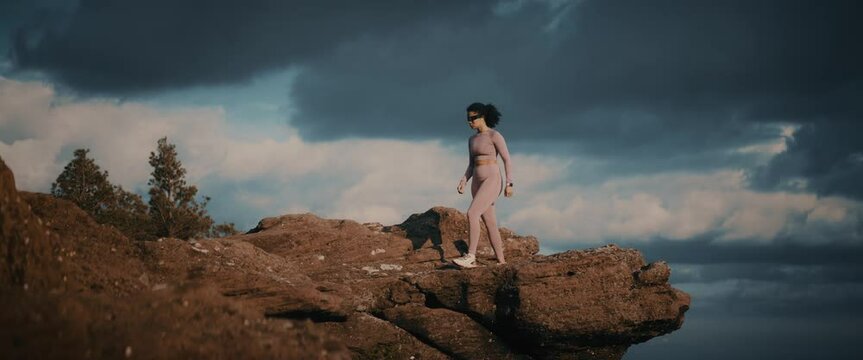 Panoramic footage of a sporty young woman in sportswear walks on a mountain rocky cliff at sunrise