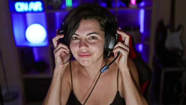 Confident young hispanic woman, beautiful streamer, engrossed in game, streaming in dark gaming room, technology glowing, wearing headphones, ebullient smile lighting up the night