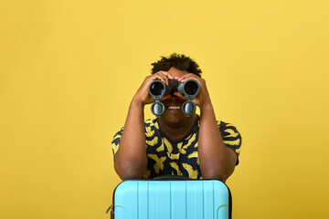 Funny young black man on a yellow background with a suitcase watching through modern binoculars. An...