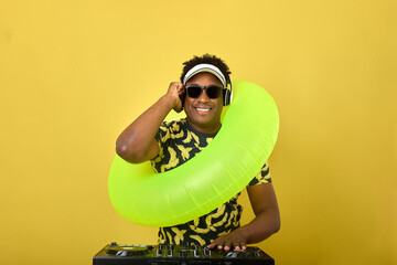 Fashionable modern dark-skinned guy DJ put on an inflatable circle, plays popular music. Cheerful handsome man in headphones and sunglasses posing at the DJ console on a yellow background