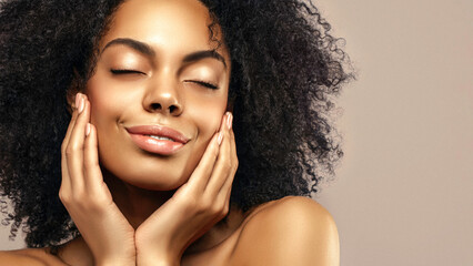 African American skincare model with a glowing healthy skin and curly hair is smiling and posing with a closed eyes against beige background. Beauty spa care treatment concept.