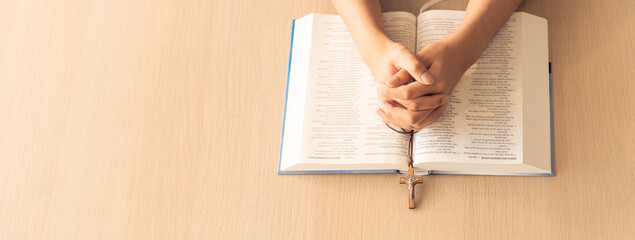 Fototapeta na wymiar Cropped image of praying male hand holding cross on holy bible book at wooden table. Top view. Concept of hope, religion, faith, christianity and god blessing. Warm and brown background Burgeoning.