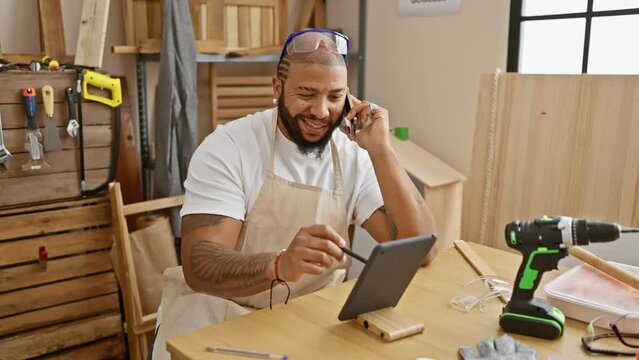 Smiling man with beard using tablet and talking on phone in a woodwork workshop.