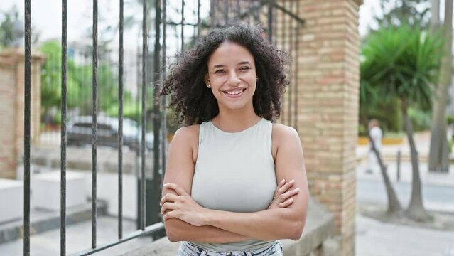 Joyful young hispanic woman flaunting her beautiful curly hair and infectious smile, standing outside in the heart of the city, radiating positivity with her arms crossed in a confident stance