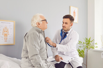 Obraz premium Male young doctor with stethoscope examining senior elderly man's lungs, breathing and heartbeat sitting on couch in exam room during medical checkup in clinic. Health care and medicine concept.