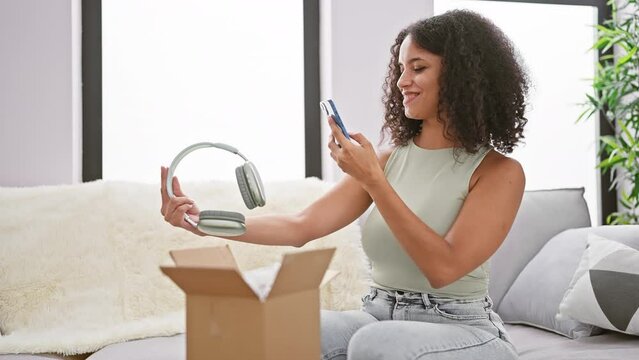 Exciting unboxing, confident, smiling young hispanic woman with curly hair, sitting at home, taking a picture of her new headphones with her phone in a cozy living room