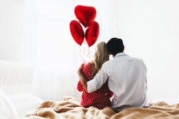Couple. Love. Valentine's day. Back view of man and woman sitting on the bed, she is holding red heart-shaped balloons
