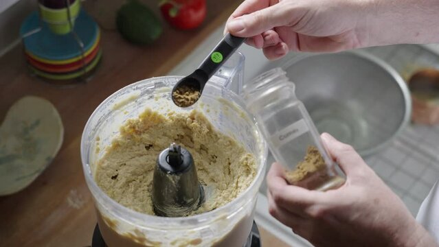 Adding cumin spice to chickpeas, hummus in a food processor, using a measuring spoon, in a farmhouse kitchen, in natural light