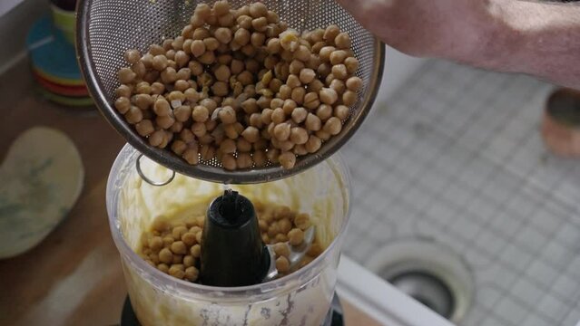Adding chickpeas to the contents of a food processor, using a metal strainer, in a farmhouse kitchen, in natural light