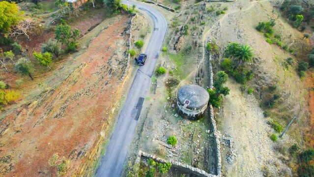 aerial drone shot focussing on blue SUV car flying away on small narrow rural village road in rayta hills udaipur showing the desert landscape of this tourist spot