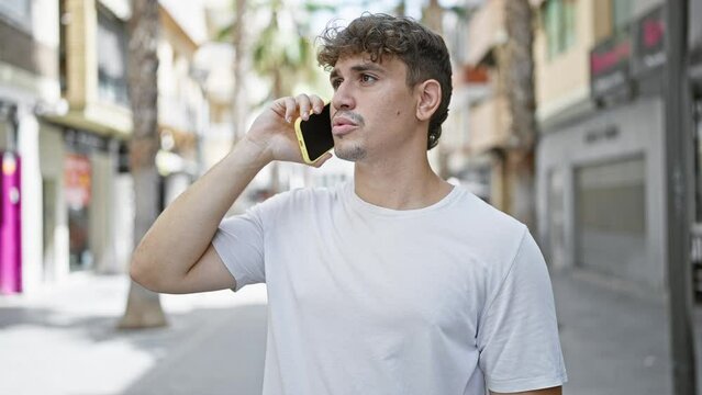 Cool-looking, handsome young hispanic man seriously engaged in conversation on his smartphone, standing under the sunny urban street's casual fashion backdrop with a concentrated expression.