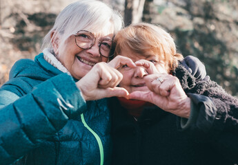 A smiling couple of elderly women hug each other in the park forming a heart with their hands looking at the camera, two elderly friends or family members are together expressing love and tenderness