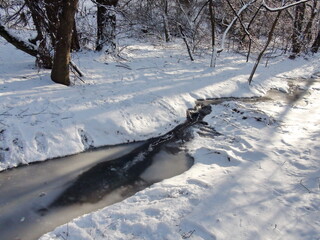The landscape of a forest river that with difficulty breaks through the ice cover surrounded by snow-white banks under the rays of the frosty sun.
