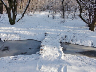 A traveler's view of a shaky bridge over a small forest river covered with an ice shell.