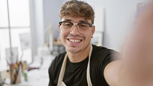 Confident young hispanic man artist, sitting, smiling brightly during a video call, talking passionately about his painting process indoors at the studio