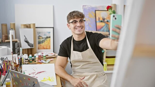 Attractive young hispanic man artist, smiling while taking self-portrait selfie with smartphone at indoor art studio, surrounded by paintbrushes, canvas and the aroma of fresh paint.