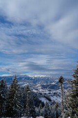 Snow-covered mountain range, against the background of blue sky and clouds, viewed from the forest