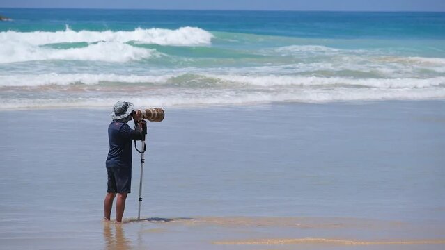 A photographer on a beautiful beach takes pictures of surfers with a camera with a large telephoto lens and a monopod