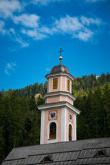 Ein pfirsichfarbener Kirchturm mit Kreuz auf der Spitze, vor einem Waldhang und blauem Himmel