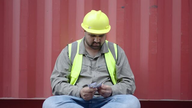 Asian container male worker or dock foreman sitting and counting the dollar money salary by his hand after working hard all month,  engineering business financial people concept, earning or profit 
