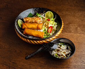 Asian cuisine. Rice noodles and fried rolls and onion sauce in black ceramic plates on a wooden table background