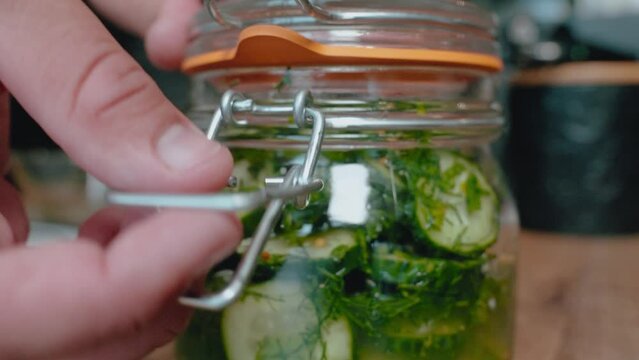 Hands are closing glass jar filled with cucumber slices dill. wooden surface with a blurry background, focusing on home canning and the preparation of pickles. This is common food preservation process