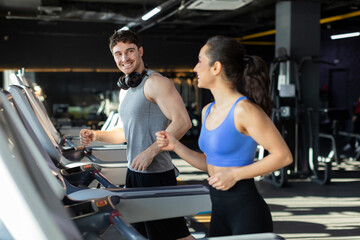 Excited young couple running on treadmills, looking at each other and smiling, enjoying cardio workout in modern gym interior, copy space - obrazy, fototapety, plakaty