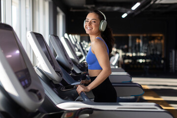 Smiling young woman happily exercises on treadmill, wearing headphones and looking at camera, radiating positive energy in a joyful fitness moment