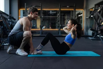 Tuinposter Young fit couple working out at gym, man helping lady doing abs exercises with ball woman and handsome muscular guy training in dark modern gym © Home-stock