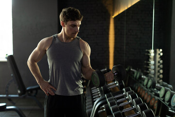 Naklejka premium Young man bodybuilder working out with dumbbells weights at the gym, standing near mirror, free space