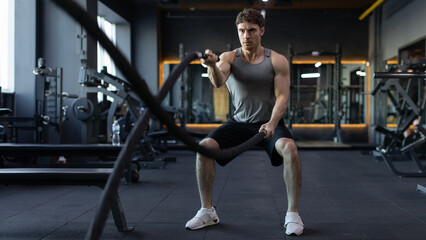 Athletic young european man with battle ropes doing exercises in functional training fitness gym, panorama