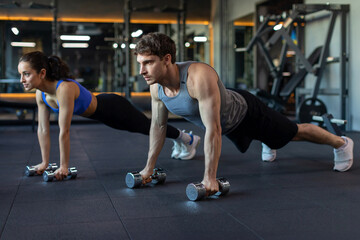 Young caucasian couple working out together, doing plank on dumbbells or push-ups on weights, training in modern gym interior