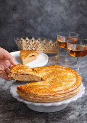A plate of traditional French galette des rois while hand grabbing its piece about to eat with crown and glases of cider in background. Cake made with puff pastry and creamy almond filling.