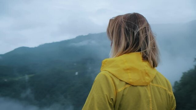 Female traveler in yellow raincoat looking from bridge in foggy mountains