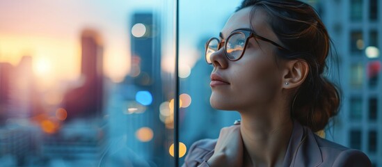 Thoughtful young businesswoman with glasses looking out office windows at city skyline.
