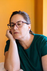 Portrait of woman in her 50s with glasses and brown hair pulled back, serious, sad and pensive with a lost gaze and hand over her face. Concept of mental health and wellness