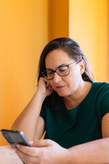Portrait of 50 years old woman with loose brown hair and glasses, looking at her cell phone with relaxed and calm attitude. Concept of technology and mature age