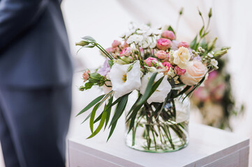 A glass vase with colorful flowers with water stands on a wooden table at a wedding ceremony.