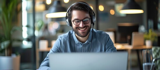 Young successful consultant working in a call center, happily using headphones to listen and take notes on audio course while typing on a laptop.