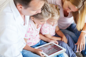 Happy family of mother, father and daughters sitting on a sofa at home having fun using a tablet...